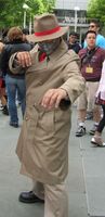 Man in beige trenchcoat and fedora (with red hatband) and silver mask